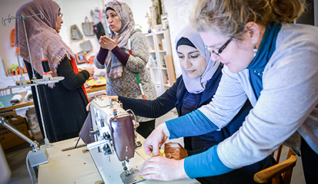 Eine Gruppe Frauen in einer Nähwerkstatt. Zwei Frauen mit Kopftuch sind im Gespräch. Eine Frau mit Locken und Brille und eine weitere Frau mit Kopftuch erbeiten an einer Nähmaschine. | Sparkasse Hannover