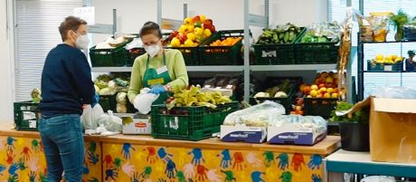 Frau mit Schürze und FFP maske gibt an einem Tresen voller Obst und Gemüse Essen an Frau in Jeans und Pullover und FFP Maske aus | Sparkasse Hannover