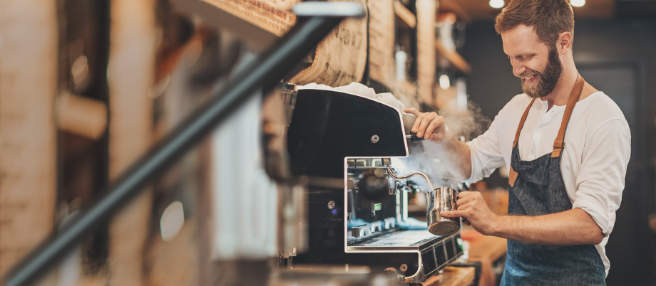 Junge Leute: Studenten - Junger Mann mit Schürze steht vor einem Kaffeeautomaten und bereitet Kaffee zu | Sparkasse Hannover