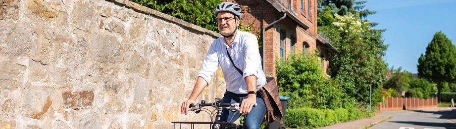 Ein Mann mit hochgekrämpelten Hemdärmeln, einer Umhängetasche und einem Fahrradhelm fährt auf einem Fahrrad | Sparkasse Hannover