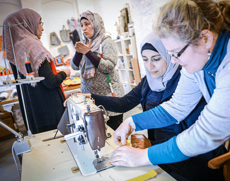Eine Gruppe Frauen in einer Nähwerkstatt. Zwei Frauen mit Kopftuch sind im Gespräch. Eine Frau mit Locken und Brille und eine weitere Frau mit Kopftuch erbeiten an einer Nähmaschine. | Sparkasse Hannover