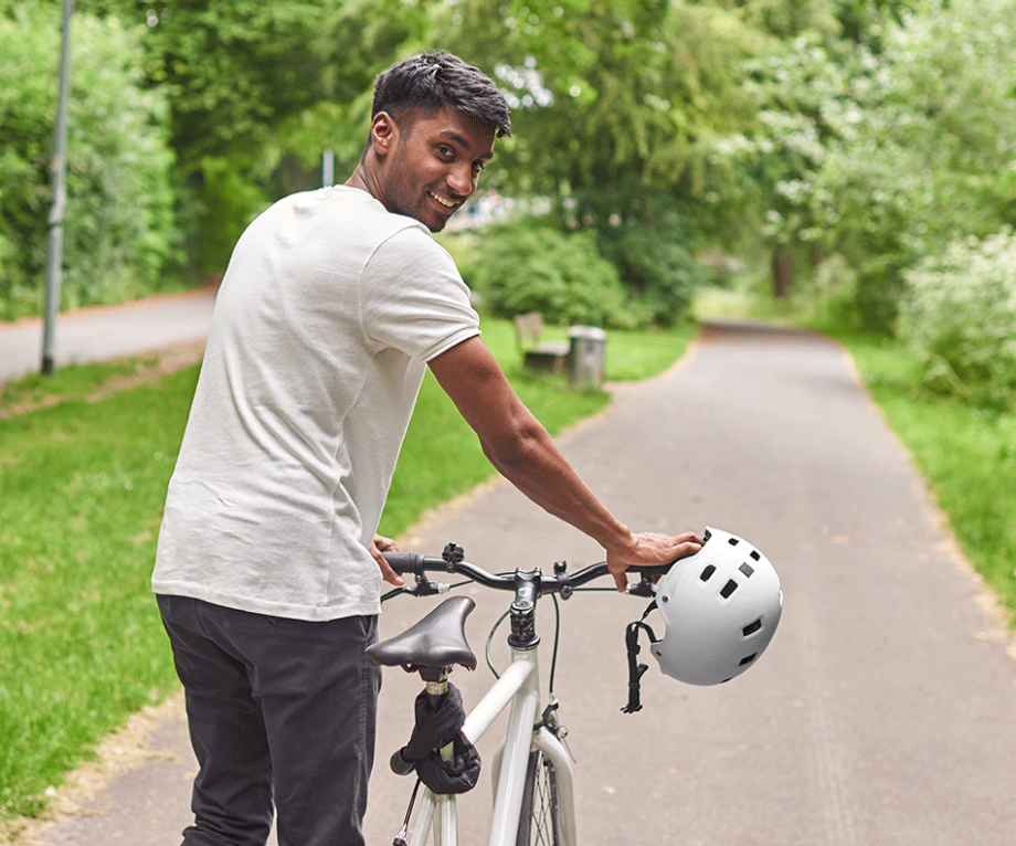 Ein junger Mann in Jeans und T-Shirt schiebt lächelnd ein Fahrrad in grüner, urbaner Umgebung| Sparkasse Hannover