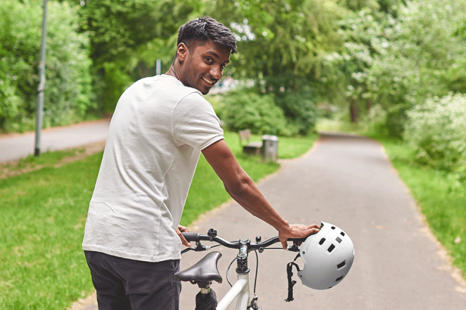 Ein junger Mann in Jeans und T-Shirt schiebt lächelnd ein Fahrrad in grüner, urbaner Umgebung| Sparkasse Hannover