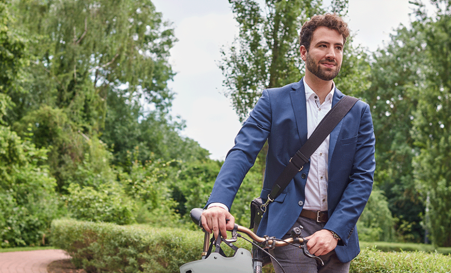 Junger Mann in Sacko und mit Umhängetasche schiebt ein Fahrrad durch urbane, aber begrünte Umgebung | Sparkasse Hannover