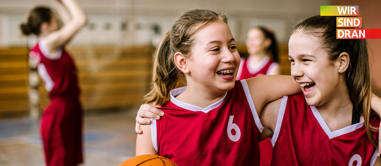 Zwei Basketball- Trickots tragende Mädchen lachen Arm in Arm und mit einem Basketball in der Hand in einer Turnhalle.| Sparkasse Hannover