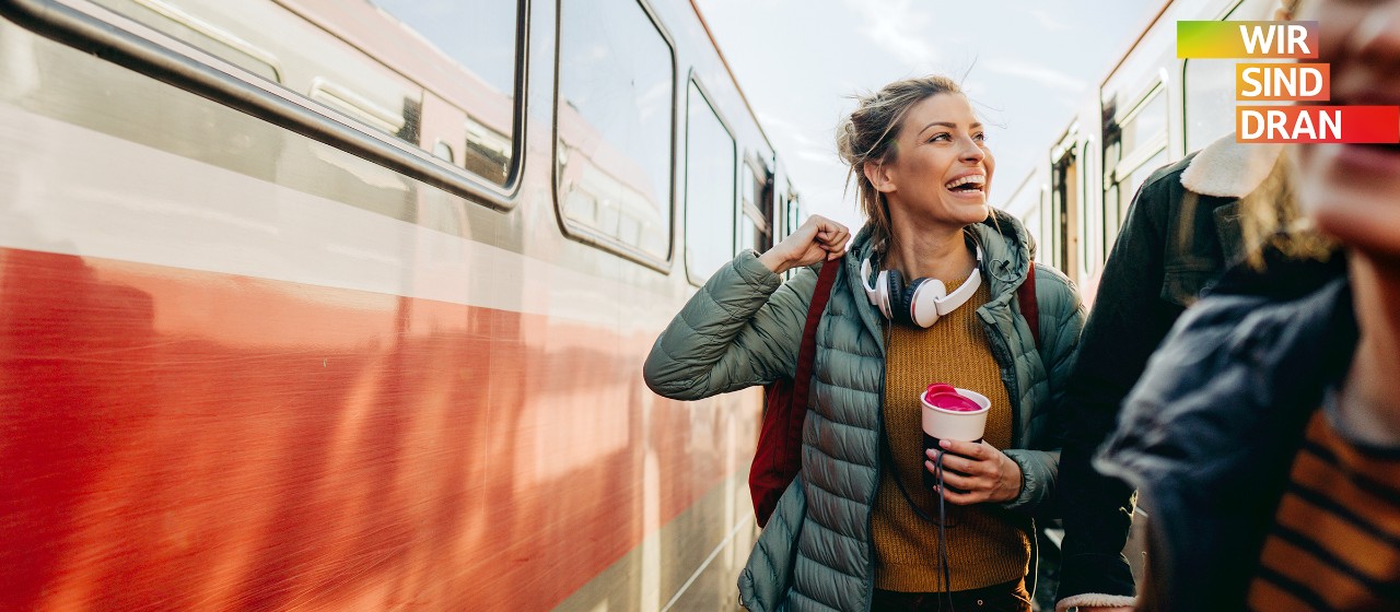 Junge Frau mit Rucksack auf dem Rücke und einem Thermosbecher in der Hand steht zwischen zwei Zügen auf einem Gleis und lacht jemanden fröhlich an | Sparkasse Hannover