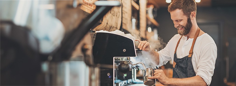 Junge Leute: Studenten - Junger Mann mit Schürze steht vor einem Kaffeeautomaten und bereitet Kaffee zu | Sparkasse Hannover