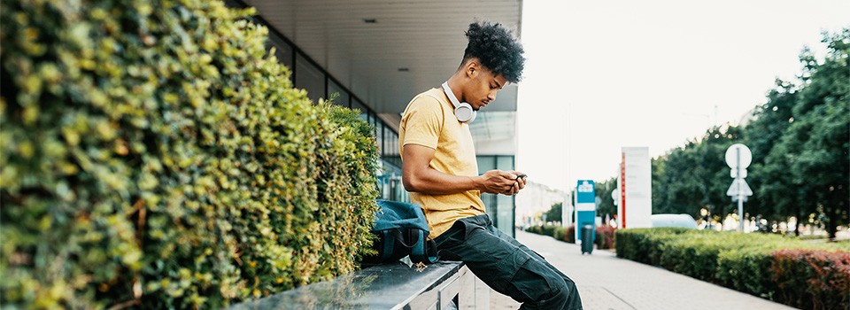 Junger Mann mit schwarzen Locken sitzt mit einem Handy in der Hand auf einer Steinmauer  | Sparkasse Hannover